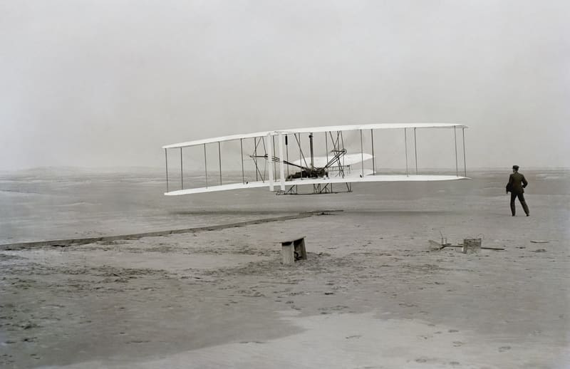 Black-and-white photograph of the Wright Flyer biplane just after takeoff, with one brother running alongside on the sand