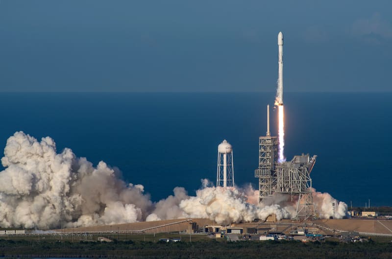 A white Falcon 9 rocket rising from the launch pad on a column of bright flame and smoke